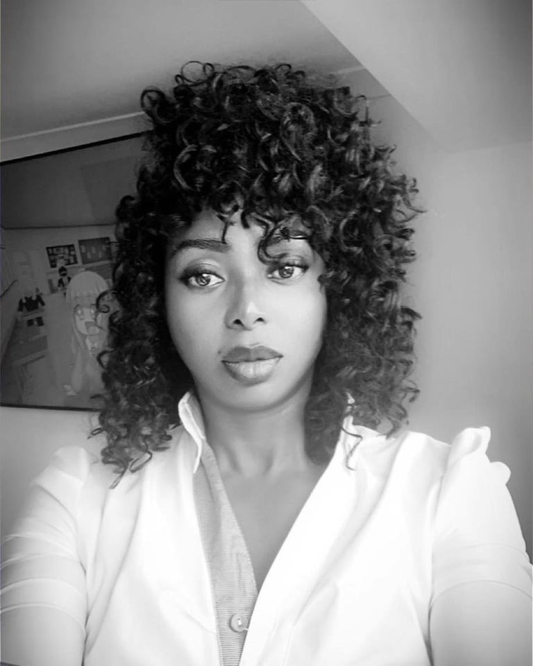 Black and white portrait of a woman with voluminous curly hair wearing a white button-up shirt, looking at camera indoors