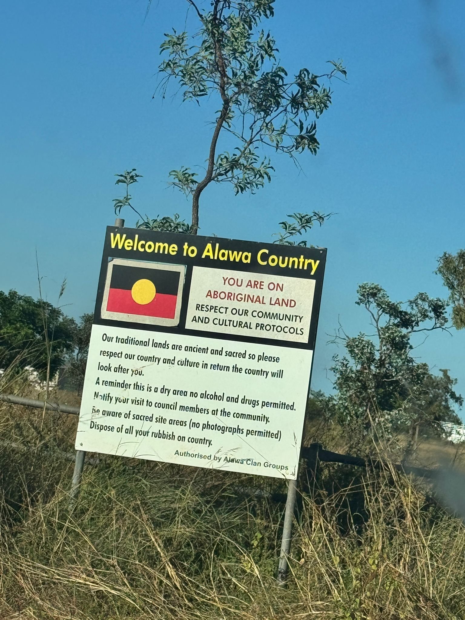 Welcome to Alawa County sign on Aboriginal land with yellow and red flag symbol against blue sky and dry grass