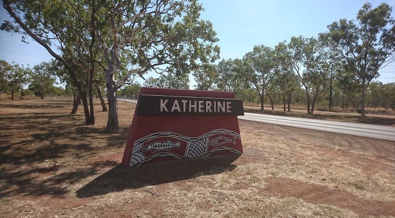 Red welcome sign for Katherine town surrounded by eucalyptus trees and red dirt road in outback Australia