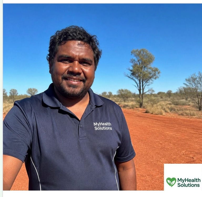 Man in navy MyHealth Solutions polo shirt smiling outdoors with red dirt and sparse trees under blue sky