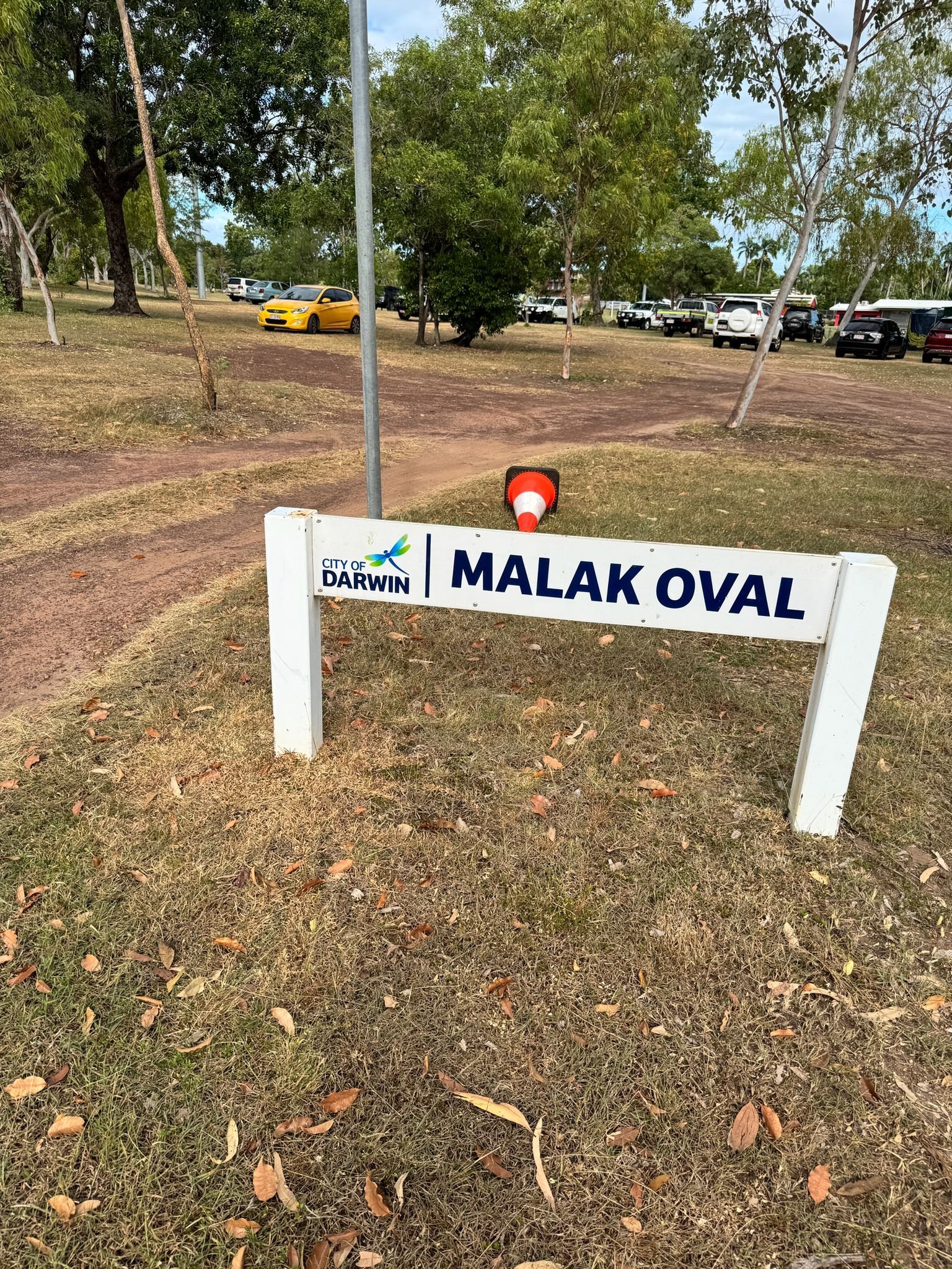 White signage for Malak Oval park with Darwin logo, red beacon light, and parking area with vehicles and trees in background
