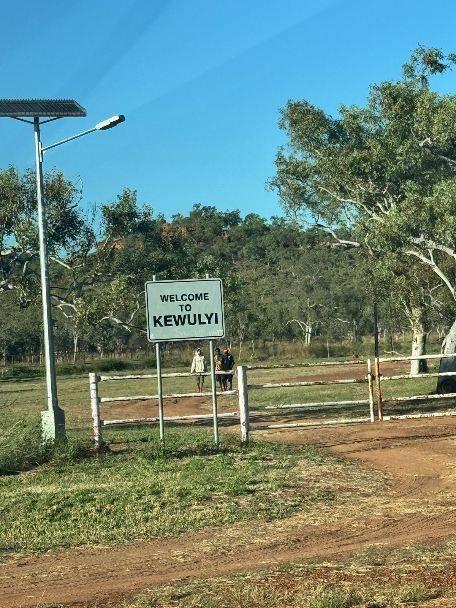 Welcome sign to Kewulyi beside a dirt road with solar lamp post and eucalyptus trees under blue sky