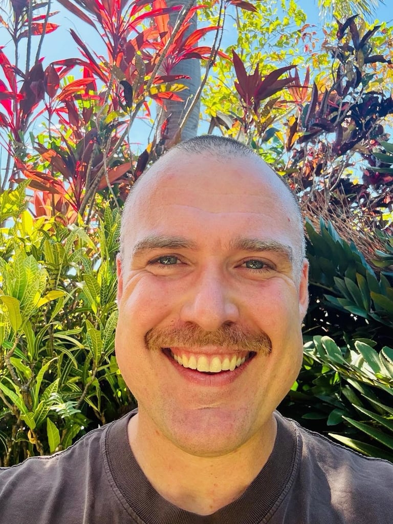 Smiling man with shaved head wearing dark shirt, posed outdoors with colorful red and green foliage in background