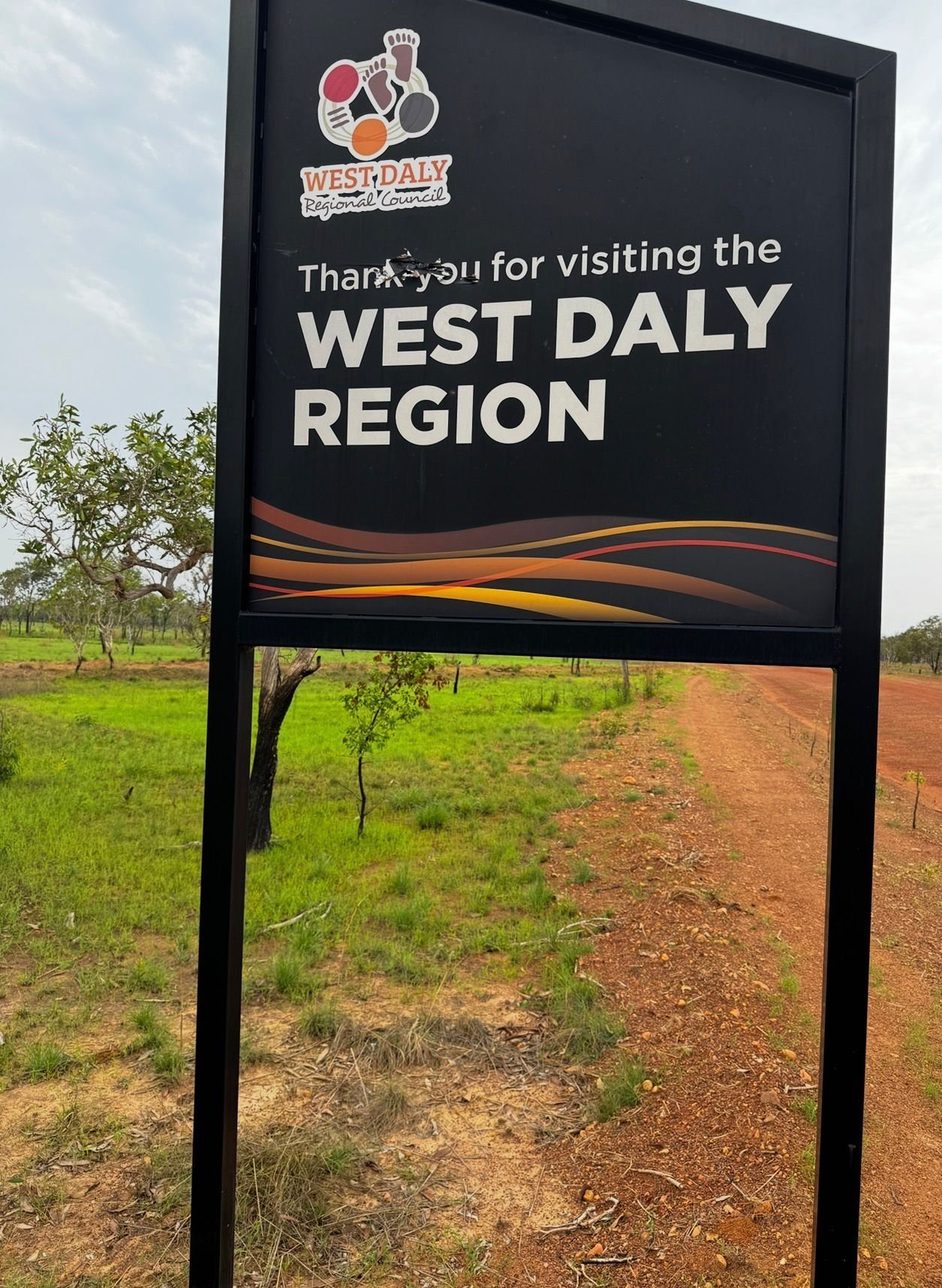 A black roadside sign thanking visitors to the West Daly Region, with the West Daly logo and red dirt landscape visible behind it