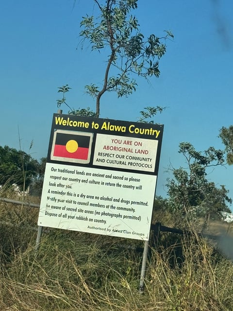 Welcome sign to Alawa County on Aboriginal land with yellow sun symbol and guidelines for visitors, surrounded by dry grass and trees under blue sky