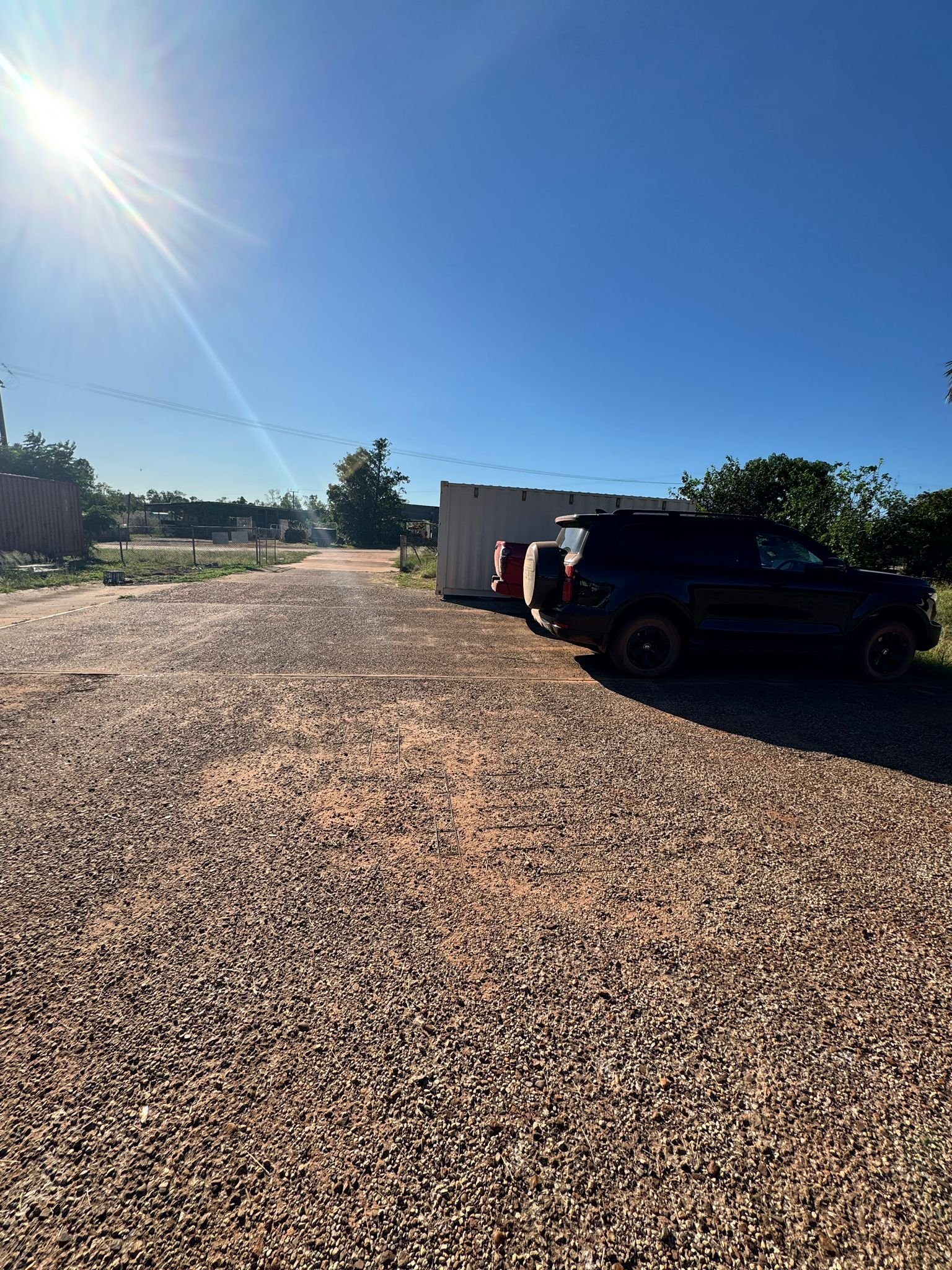 Dark SUV parked next to metal industrial buildings on a gravel lot under sunny clear blue sky