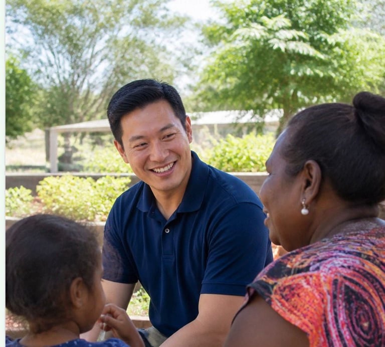 A smiling man in a blue polo shirt interacts with a woman and child in an outdoor garden setting with green trees in the background.